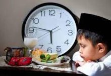 Seorang anak kecil sedang duduk di meja makan dengan tangan terlipat, menunggu waktu berbuka puasa. Latar belakang gambar adalah ruangan yang tenang dengan lampu di atas meja makan dan perabotan rumah tangga di sekitarnya.